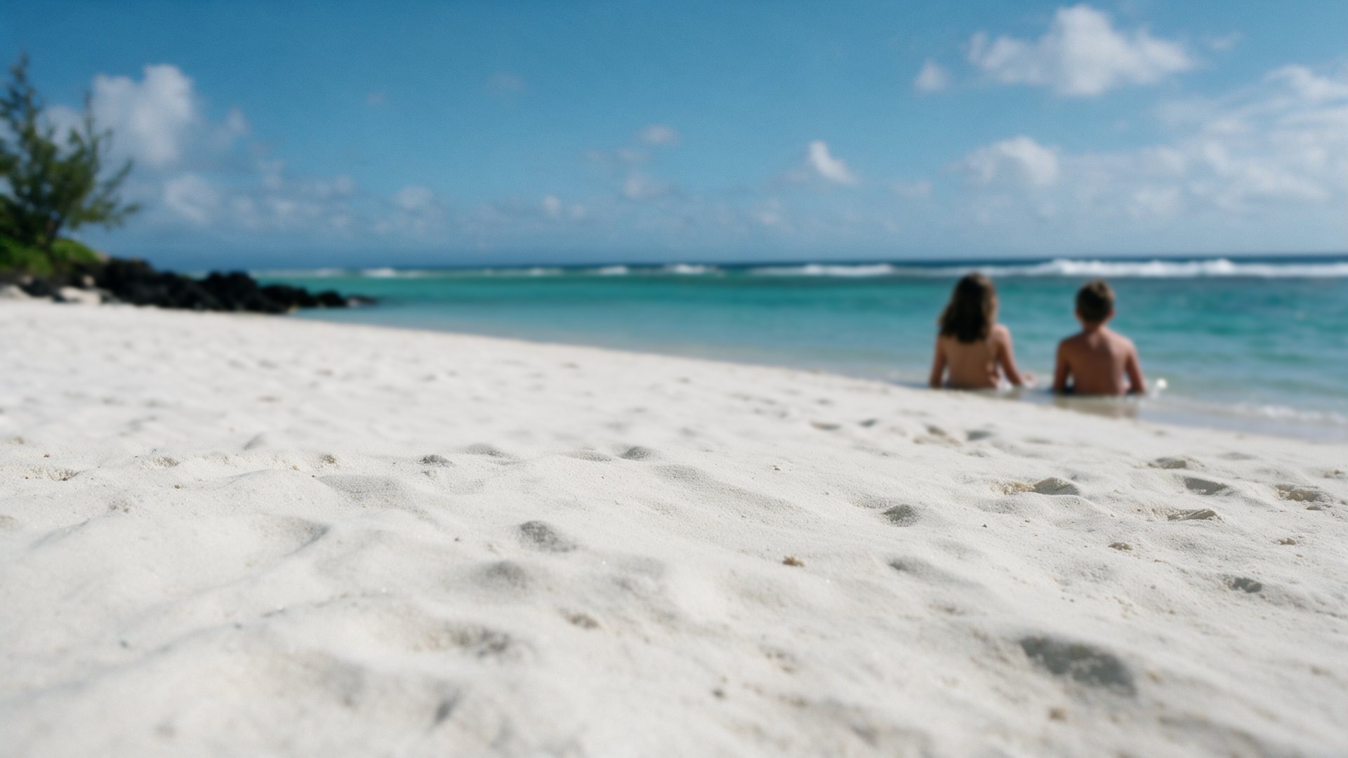 A white beach in Mauritius. Further out, two children sit in the shallow water with their backs to the camera.